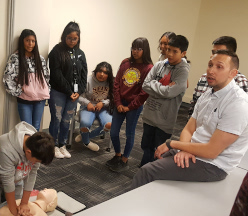 एक छात्र को नकली रोगी पर सीपीआर करते देख रहे छात्रों का समूह।
