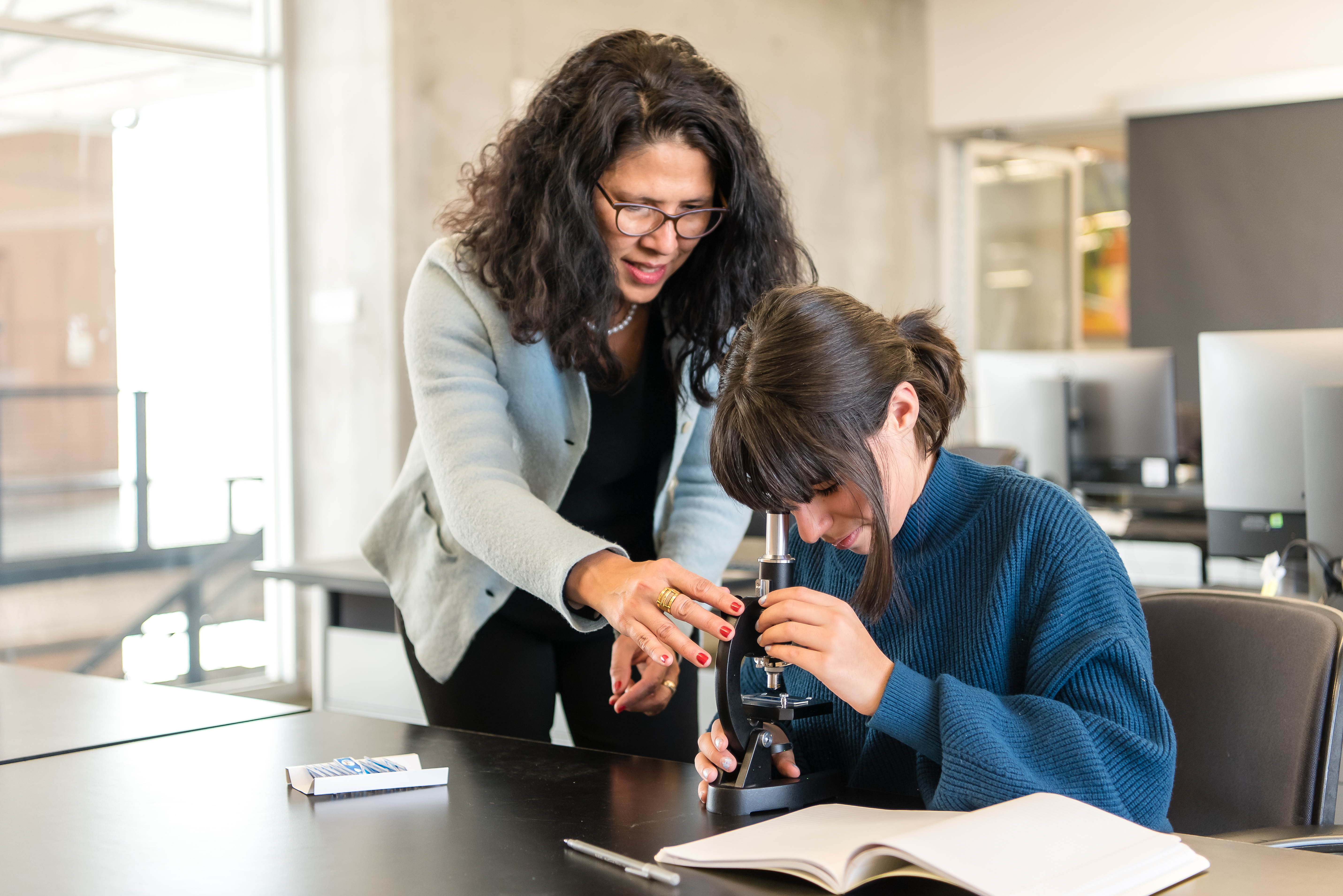 A student looks into a microscope; a teacher stands next to her