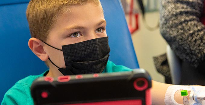 A young child in a mask looking at a tablet while their getting an infusion