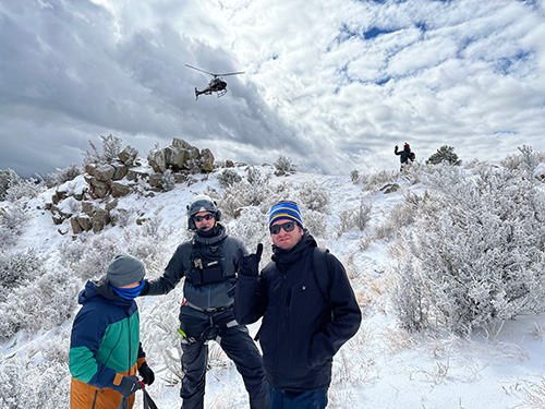 Group standing in front of helicopter flying off mountain.